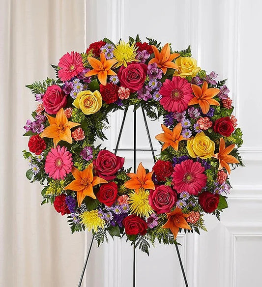 Vibrant sympathy wreath with orange lilies, red and yellow roses, gerbera daisies, carnations, and colorful accents on an easel stand.